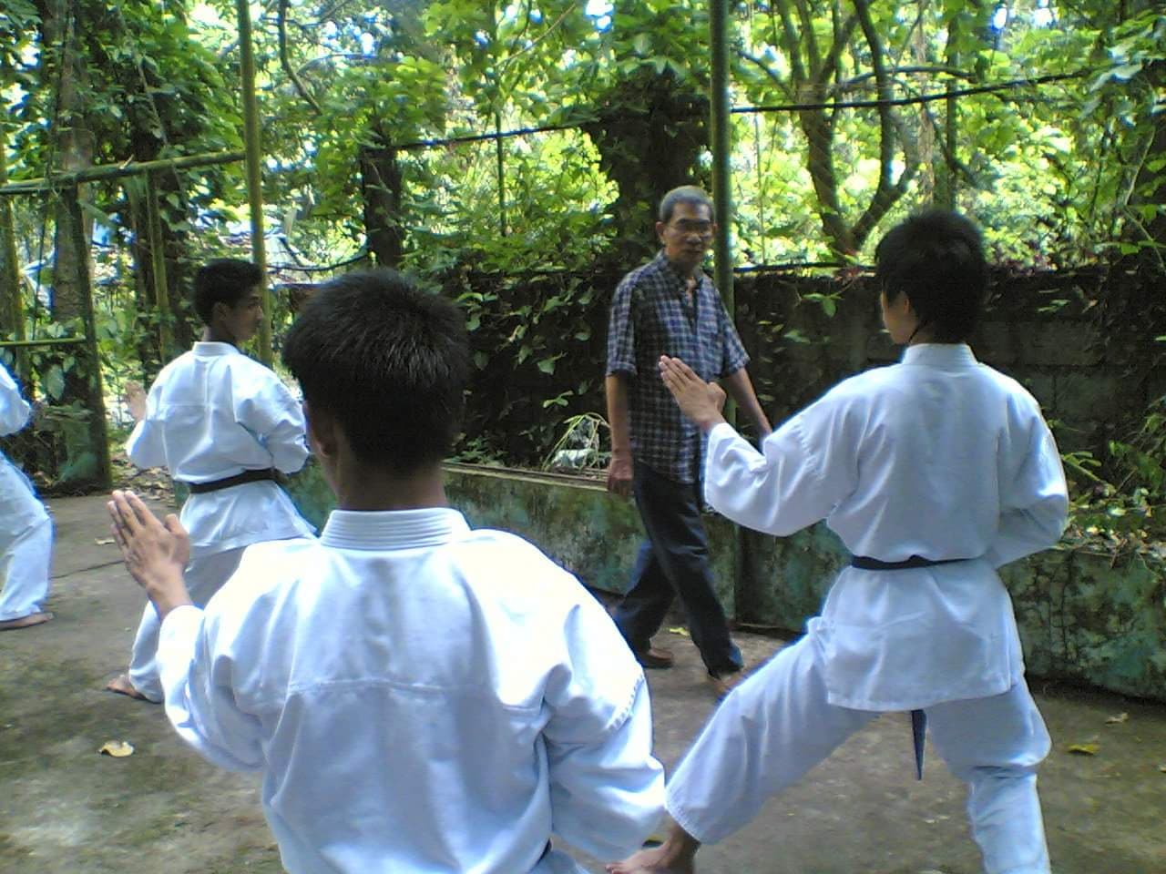 Three practitioners in white gis perform a kata on an outdoor concrete surface. Shihan Rodolfo Salinas, dressed in a checkered shirt and dark trousers, observes the practitioners from the center of the frame.